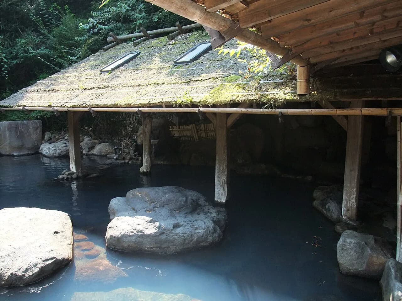 Een buitenbad van een onsen met melkblauw bronwater, grote rotsblokken en een houten afdak met mos in Kurokawa Onsen op Kyushu