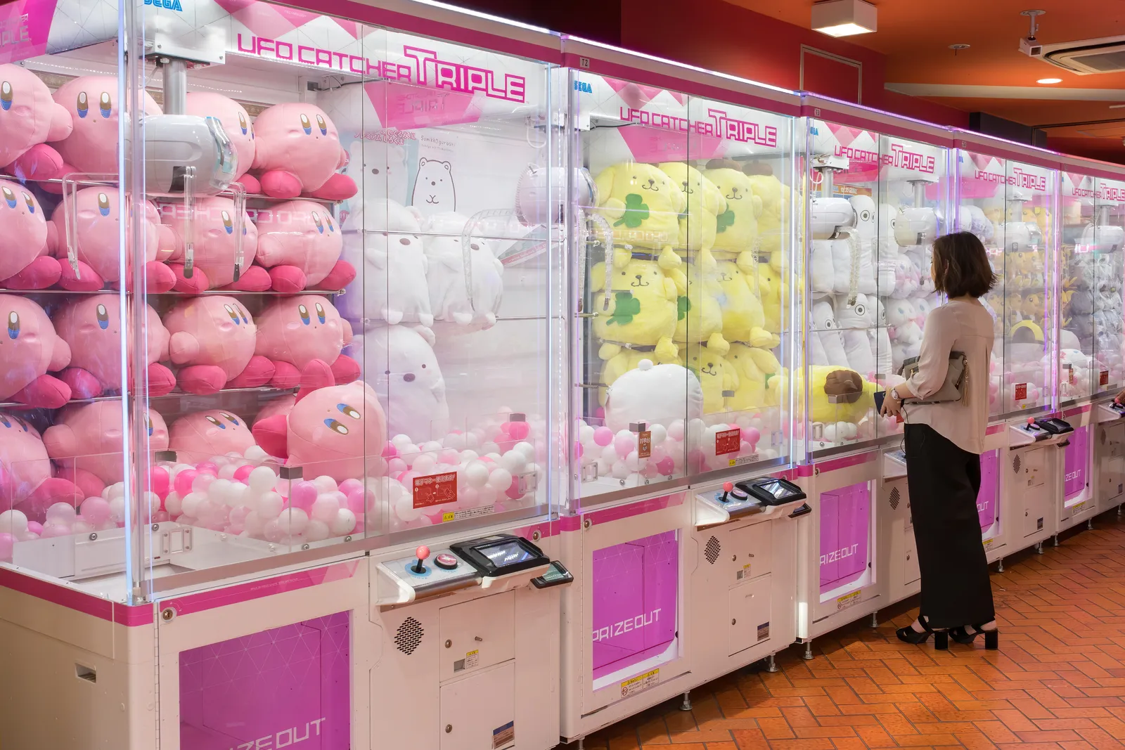 Een rij grijpautomaten in een speelhal in Akihabara vol roze, witte en gele knuffels, met rechts een vrouw die speelt